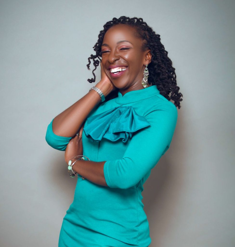 A Black woman with curly hair is smiling, wearing a teal dress with a ruffled collar. She poses with one hand on her cheek and the other arm crossed. The background is light gray.