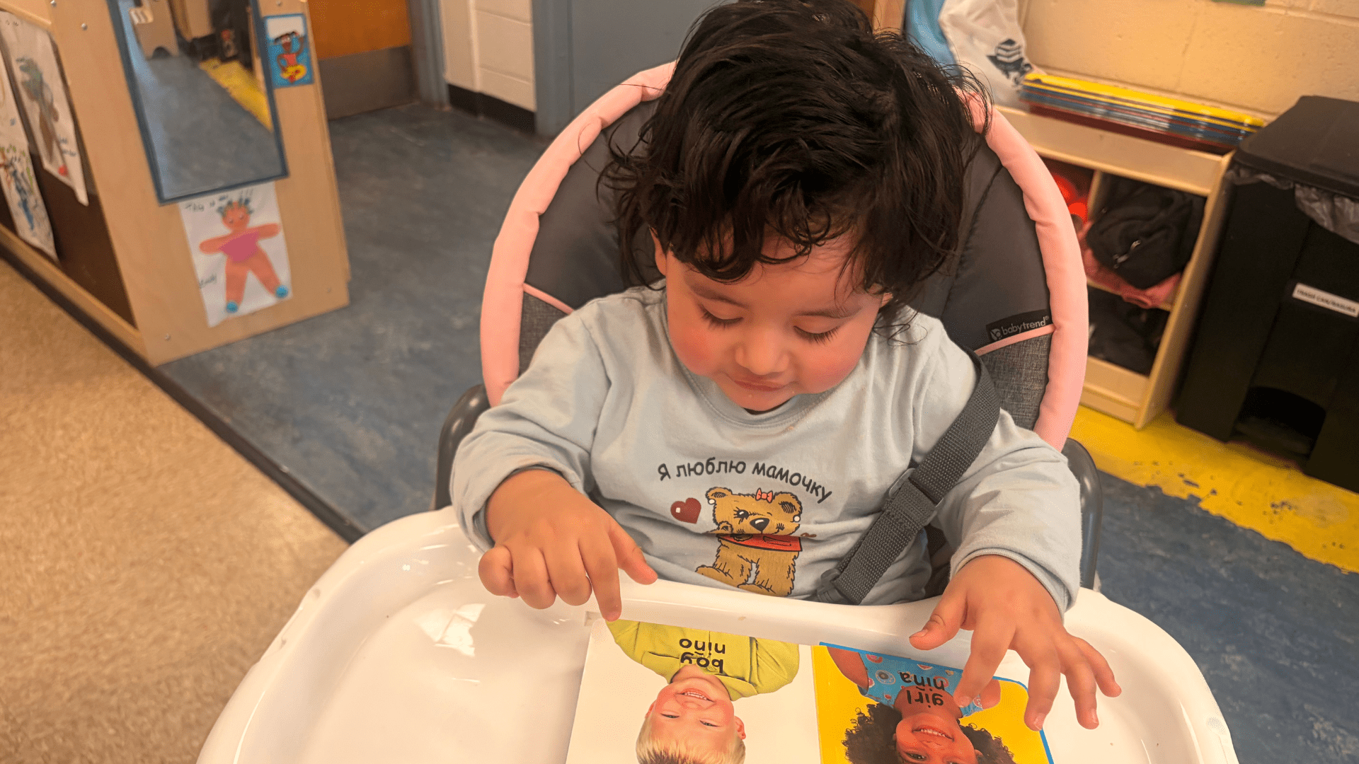 A toddler sitting in a high chair, looking at a colorful picture book featuring images of children. The background shows a classroom environment.