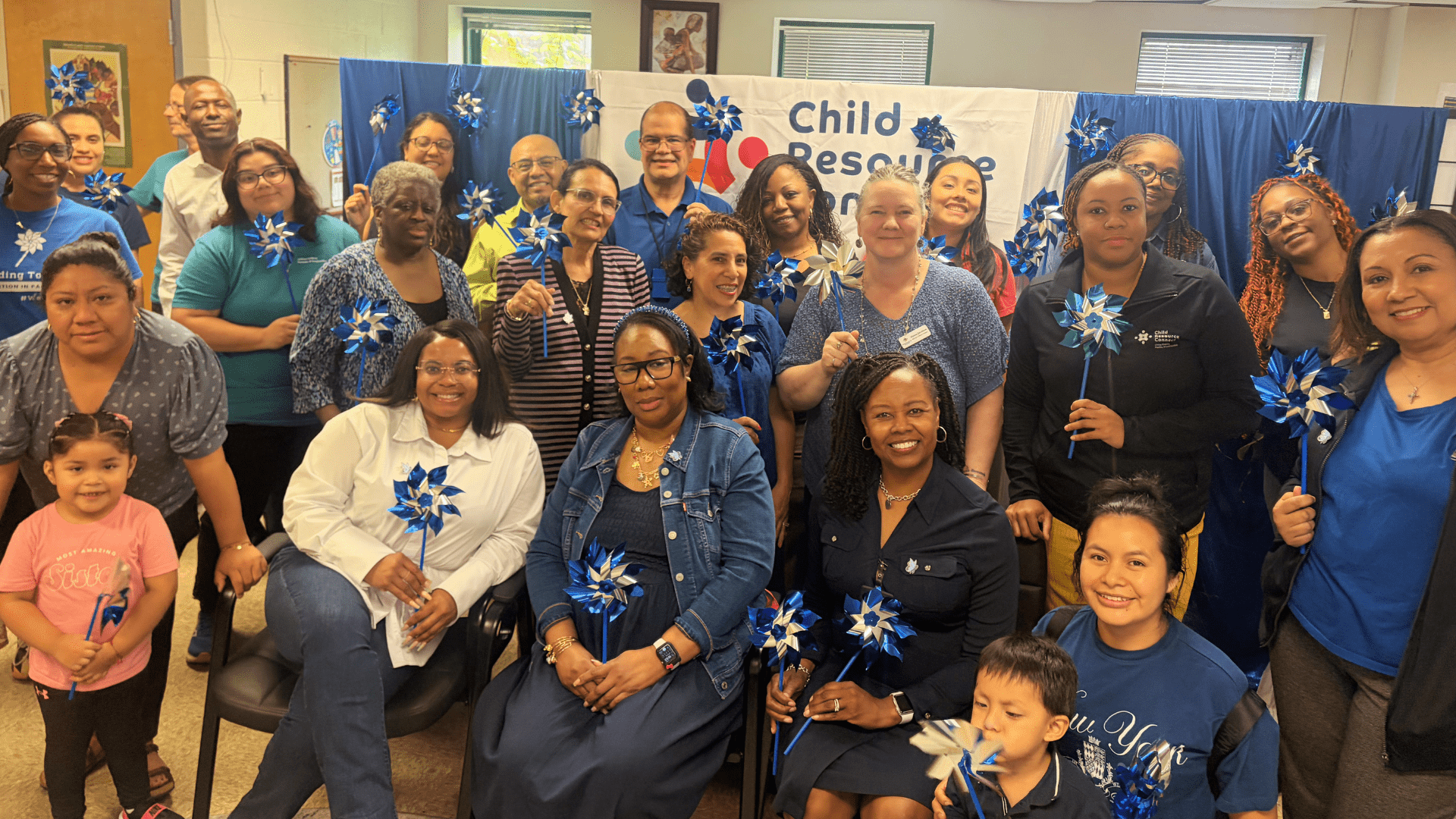 A large group of diverse adults and children pose together, holding blue pinwheel decorations, in front of a banner that reads 